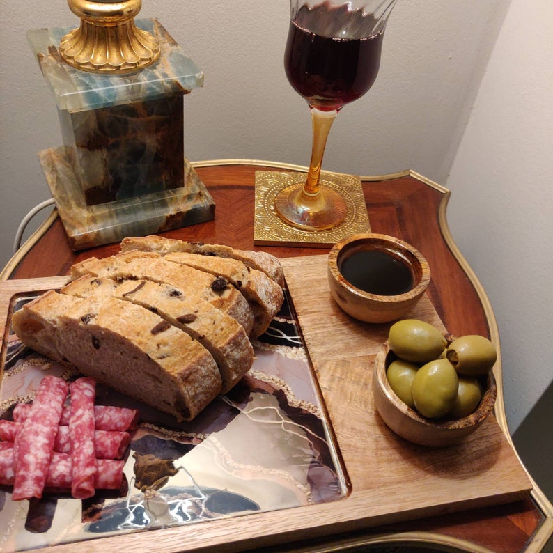 May include: A wooden serving tray with a glass insert featuring a marbled design. The tray has a small bowl of olives, a small bowl of balsamic vinegar, and a plate of sliced bread and salami.