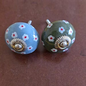 Puede incluir: Dos pomos de caj&oacute;n de cer&aacute;mica con un dise&ntilde;o floral. Un pomo es azul con flores blancas y centros rojos. El otro pomo es verde con flores blancas y centros rojos.