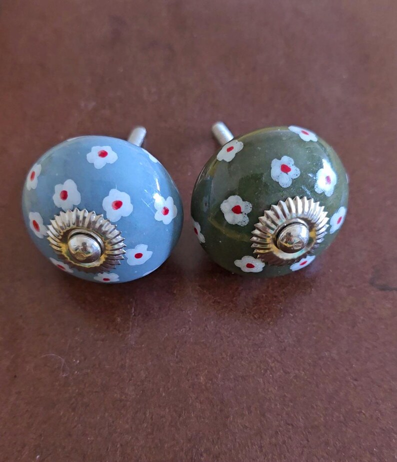 Puede incluir: Dos pomos de caj&oacute;n de cer&aacute;mica con un dise&ntilde;o floral. Un pomo es azul con flores blancas y centros rojos. El otro pomo es verde con flores blancas y centros rojos.