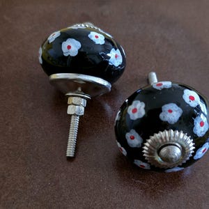 Puede incluir: Dos pomos de cer&aacute;mica negra con flores blancas y rojas. Los pomos tienen una base plateada y un tornillo para sujetarlos a un caj&oacute;n o armario.
