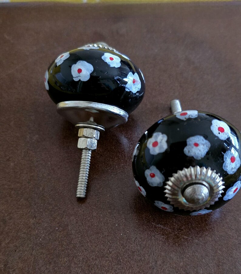 Puede incluir: Dos pomos de cer&aacute;mica negra con flores blancas y rojas. Los pomos tienen una base plateada y un tornillo para sujetarlos a un caj&oacute;n o armario.