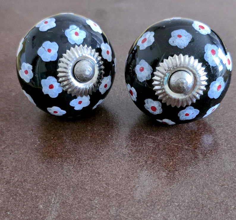 Puede incluir: Dos pomos de cer&aacute;mica negra con dise&ntilde;os florales blancos y rojos. Cada pomo tiene una base de metal plateado.