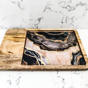 May include: A wooden serving tray with a removable insert featuring a black, pink, and gold abstract design. The design resembles agate or marble with gold glitter accents.