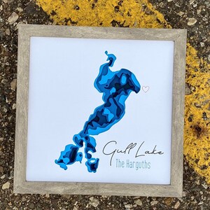 Puede incluir: Una impresión enmarcada de un lago azul con un estilo de corte de papel. El lago está rodeado de un fondo blanco y el texto "Gulf Lake The Harguths" está impreso debajo del lago. Un pequeño corazón rojo se encuentra en la esquina superior derecha de la imagen.