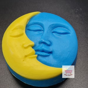 May include: A round soap bar with a celestial design. The soap is split into two sections: a yellow crescent moon and a blue face with closed eyes. The soap has a smooth texture and a detailed, carved appearance. The Soap Heaven logo is visible.