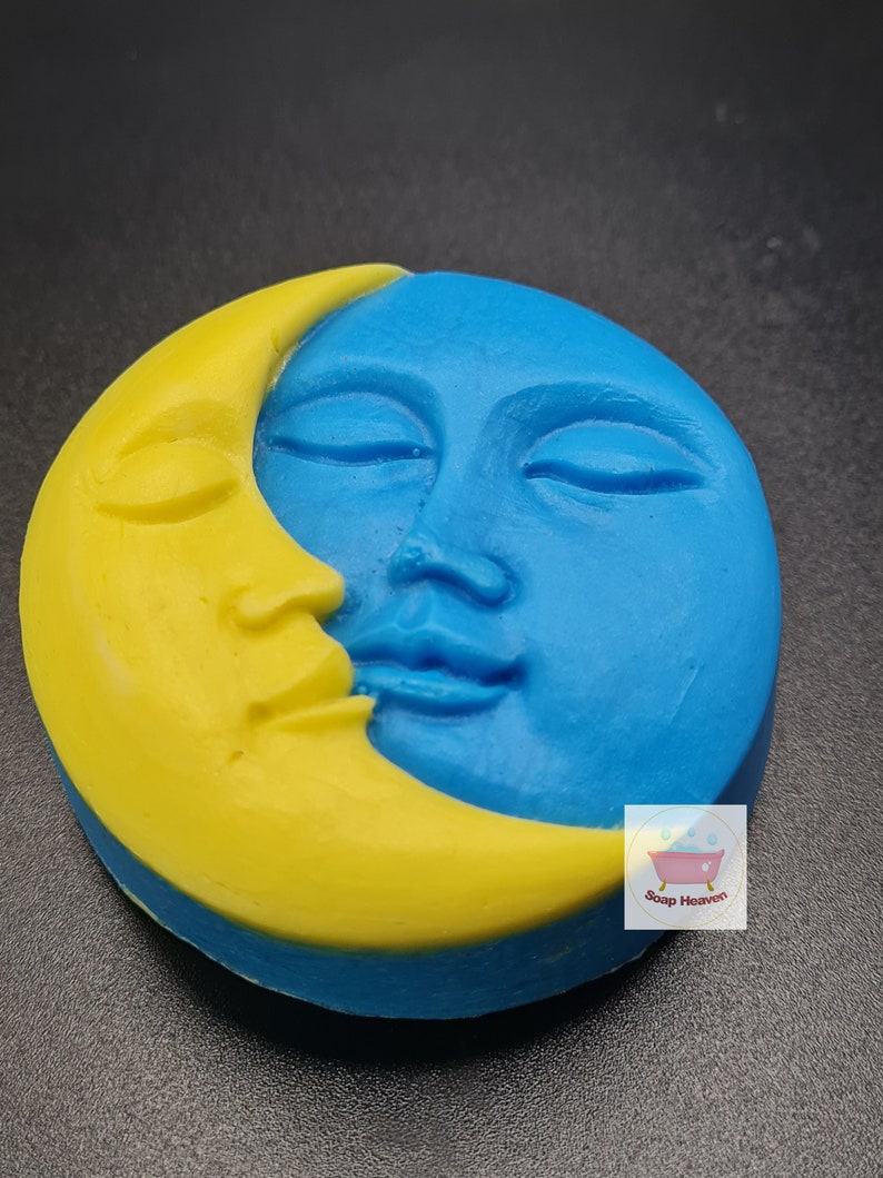 May include: A round soap bar with a celestial design. The soap is split into two sections: a yellow crescent moon and a blue face with closed eyes. The soap has a smooth texture and a detailed, carved appearance. The Soap Heaven logo is visible.