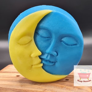 May include: A round soap bar with a celestial design. A yellow crescent moon embraces a blue face of the moon, both with closed eyes. The soap is on a wooden surface. The Soap Heaven logo is in the bottom right corner.