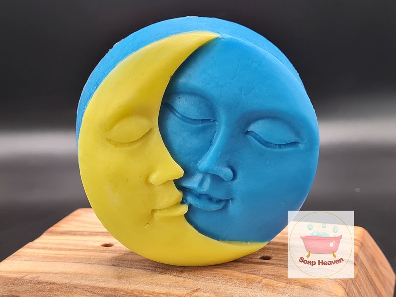 May include: A round soap bar with a celestial design. A yellow crescent moon embraces a blue face of the moon, both with closed eyes. The soap is on a wooden surface. The Soap Heaven logo is in the bottom right corner.