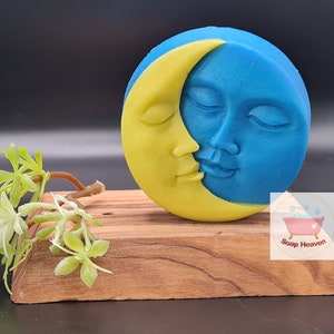 May include: A round, two-tone soap with a celestial design. The soap features a yellow crescent moon embracing a blue face with closed eyes. The soap rests on a small wooden stand, with a sprig of greenery to the left.