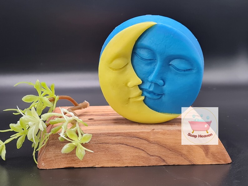 May include: A round, two-tone soap with a celestial design. The soap features a yellow crescent moon embracing a blue face with closed eyes. The soap rests on a small wooden stand, with a sprig of greenery to the left.