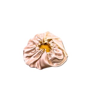 May include: A satin bonnet in a light champagne color with a golden-yellow interior. The bonnet is gathered at the edges with an elastic band, creating a ruffled effect. The fabric has a subtle sheen, reflecting light.