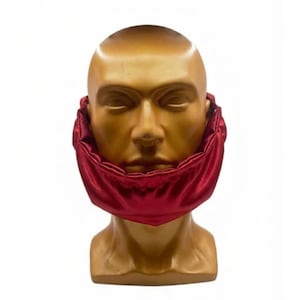 May include: A shiny, deep red satin beard cover or chin wrap positioned around the lower portion of a smooth, featureless, gold-toned mannequin head. The fabric has a gathered, elasticized edge that creates a soft, rounded, and voluminous shape.