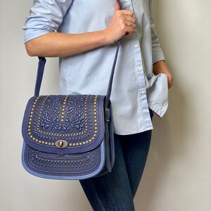 May include: A blue leather crossbody bag with intricate embossed floral designs and gold-toned accents. The bag has a flap closure with a metal buckle and a long adjustable strap.