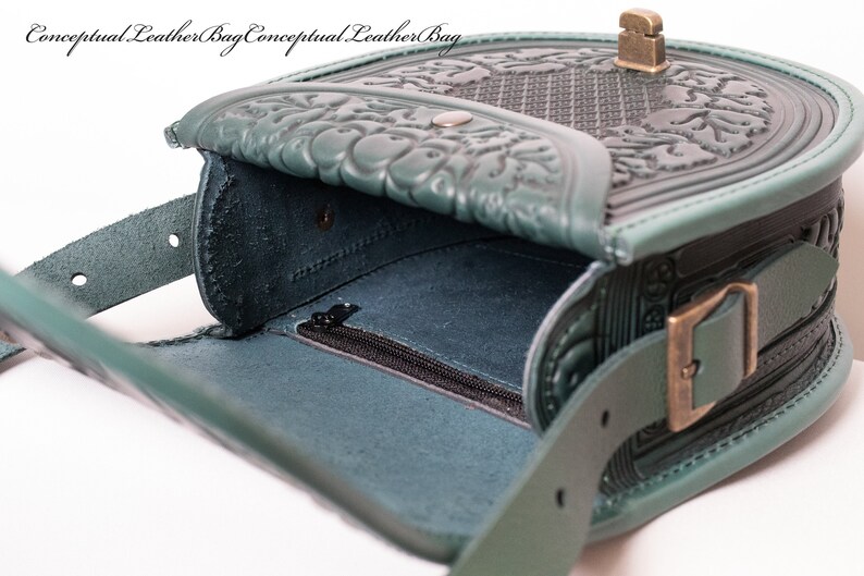 May include: A teal green leather bag with intricate embossed floral designs. The bag has a flap closure with a metal buckle and a zippered pocket on the inside.