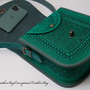 May include: A teal green leather shoulder bag with an intricate embossed design. The bag has a flap closure with a decorative metal button and a small pocket on the front. The bag is shown with a phone inside.