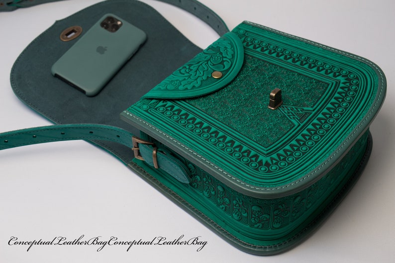 May include: A teal green leather shoulder bag with an intricate embossed design. The bag has a flap closure with a decorative metal button and a small pocket on the front. The bag is shown with a phone inside.