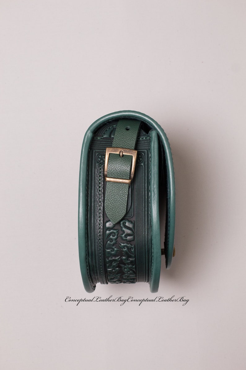 May include: A dark green leather bag with an embossed floral design and a brass buckle. The bag is closed and the strap is folded over the top.