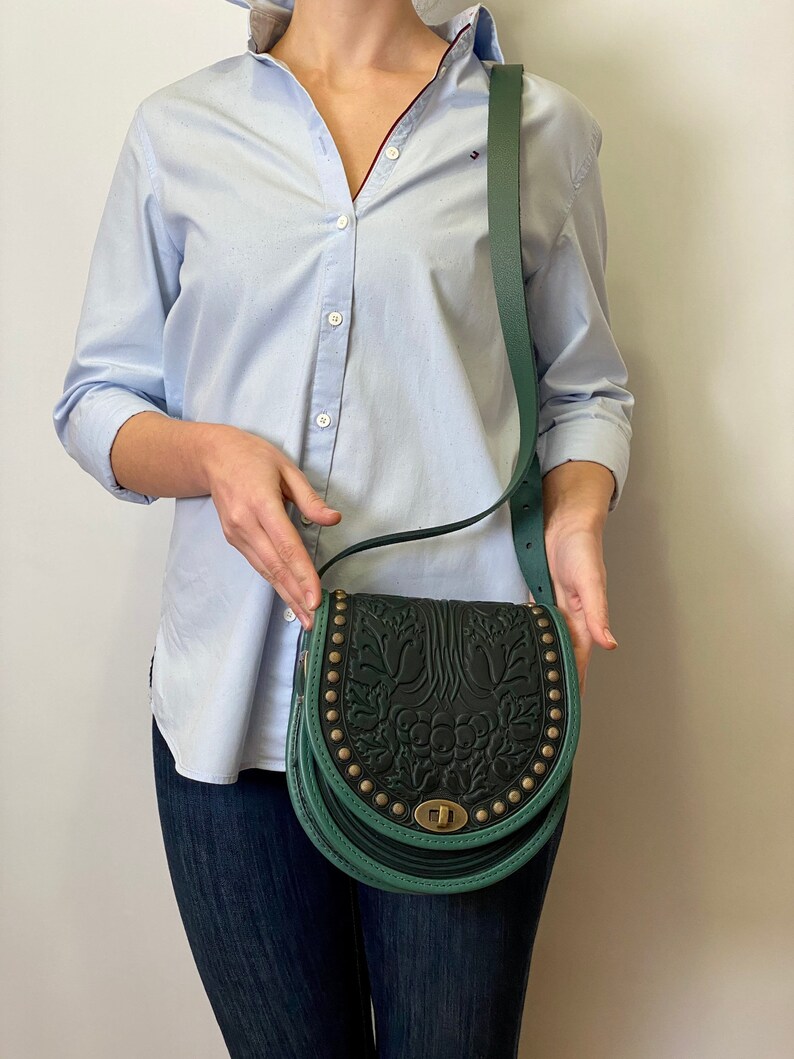 May include: A dark green leather crossbody bag with a floral embossed design and gold-toned hardware. The bag has a flap closure and a long adjustable strap.
