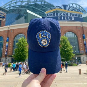 May include: A navy blue baseball cap with a gold embroidered baseball glove featuring blue flowers. The cap is held in front of American Family Field.