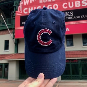 May include: A navy blue baseball cap with a red embroidered "C" on the front. The cap is held in front of a red sign that reads "Wrigley Field Home of Chicago Cubs".