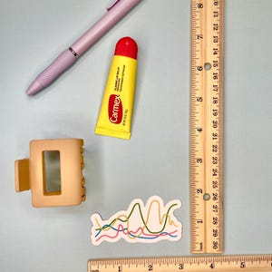 Pu&ograve; includere: Una penna a scatto viola chiaro, un tubetto di balsamo labbra Carmex giallo, un fermaglio per capelli beige opaco e un adesivo con linee ondulate colorate. Un righello di legno mostra che gli oggetti misurano circa 7-13 cm.