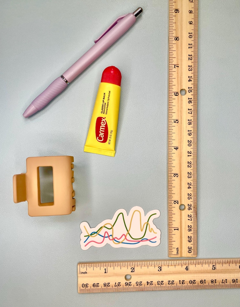 Pu&ograve; includere: Una penna a scatto viola chiaro, un tubetto di balsamo labbra Carmex giallo, un fermaglio per capelli beige opaco e un adesivo con linee ondulate colorate. Un righello di legno mostra che gli oggetti misurano circa 7-13 cm.