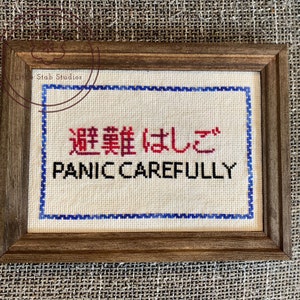 May include: A framed cross-stitch with the text "Panic Carefully" in black letters and the Japanese text "避難はしご" in red letters. The cross-stitch is framed in a dark brown wood frame.