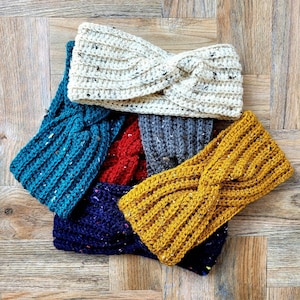 May include: Five crocheted headbands in different colors and patterns. The headbands are arranged in a pile, with the top headband being white with black flecks. The other headbands are teal, red, navy blue with white flecks, and yellow.
