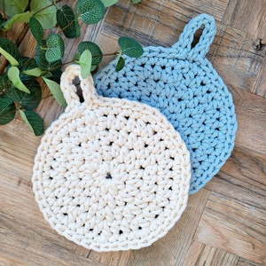 May include: Two crocheted pot holders, one in beige and one in light blue, with a loop handle on each. The pot holders are on a wooden surface.