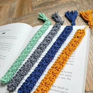 May include: Five crocheted bookmarks with tassels in different colors: mint green, gray, navy blue, mustard yellow, and a light brown. The bookmarks are lying on a book with text.