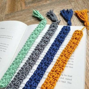 May include: Four crocheted bookmarks with tassels in different colors: green, gray, blue, and yellow. The bookmarks are laid out on a book.
