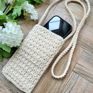 May include: A crocheted beige phone case with a long strap. The case is lying on a wooden surface with a bouquet of white flowers in the background.