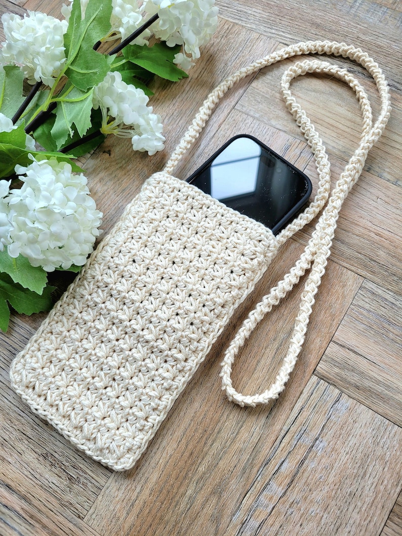 May include: A crocheted beige phone case with a long strap. The case is lying on a wooden surface with a bouquet of white flowers in the background.