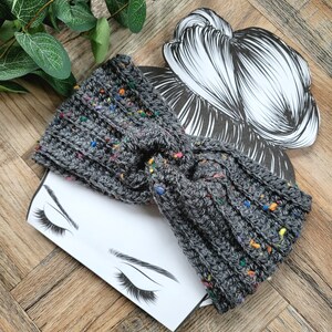 May include: Gray knitted headband with colorful flecks. The headband is twisted into a knot and is shown on a white background with a drawing of a woman's face.