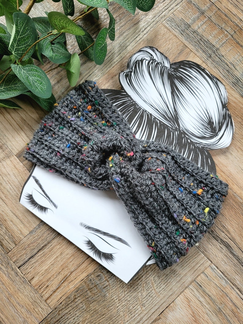 May include: Gray knitted headband with colorful flecks. The headband is twisted into a knot and is shown on a white background with a drawing of a woman's face.