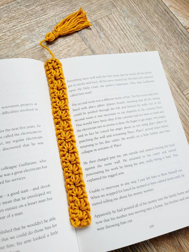 May include: A close-up of a yellow crocheted bookmark with a tassel. The bookmark is placed on a book with text about a renovation project and an electrician.