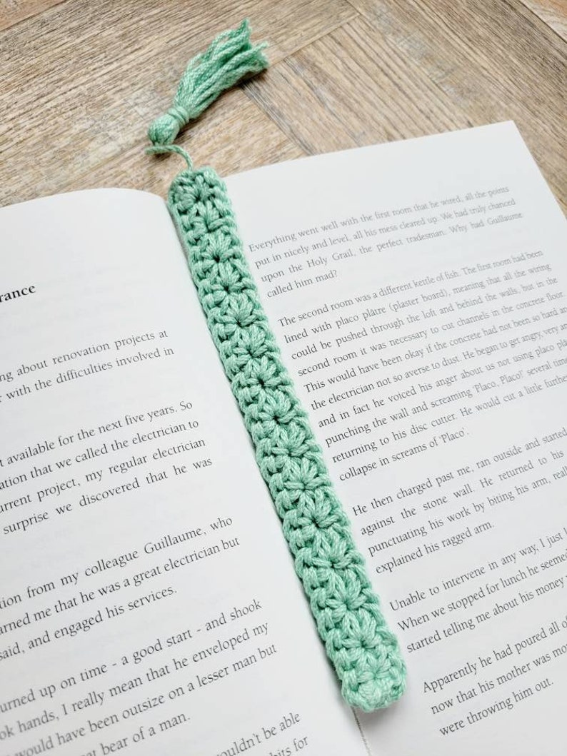 May include: A light green crocheted bookmark with a tassel. The bookmark is placed on a book with text about a renovation project.