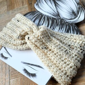 May include: A cream-colored crocheted headband with a speckled pattern. The headband is tied in a knot and is shown on a white background with a drawing of a woman's face.