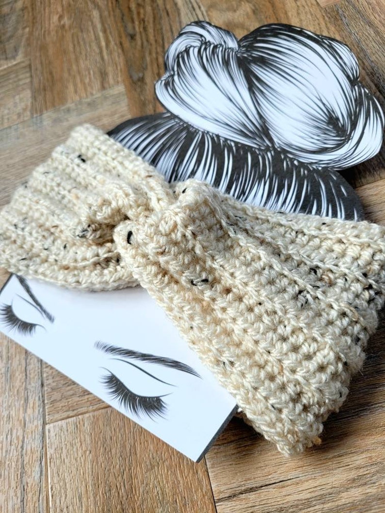 May include: A cream-colored crocheted headband with a speckled pattern. The headband is tied in a knot and is shown on a white background with a drawing of a woman's face.