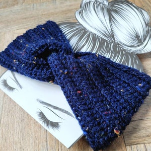 May include: A navy blue crocheted headband with flecks of red, yellow, and white yarn. The headband is twisted and knotted, creating a stylish and comfortable accessory.
