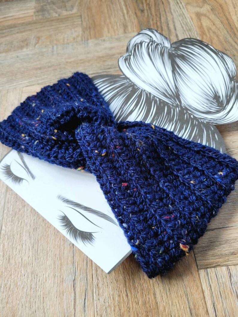 May include: A navy blue crocheted headband with flecks of red, yellow, and white yarn. The headband is twisted and knotted, creating a stylish and comfortable accessory.