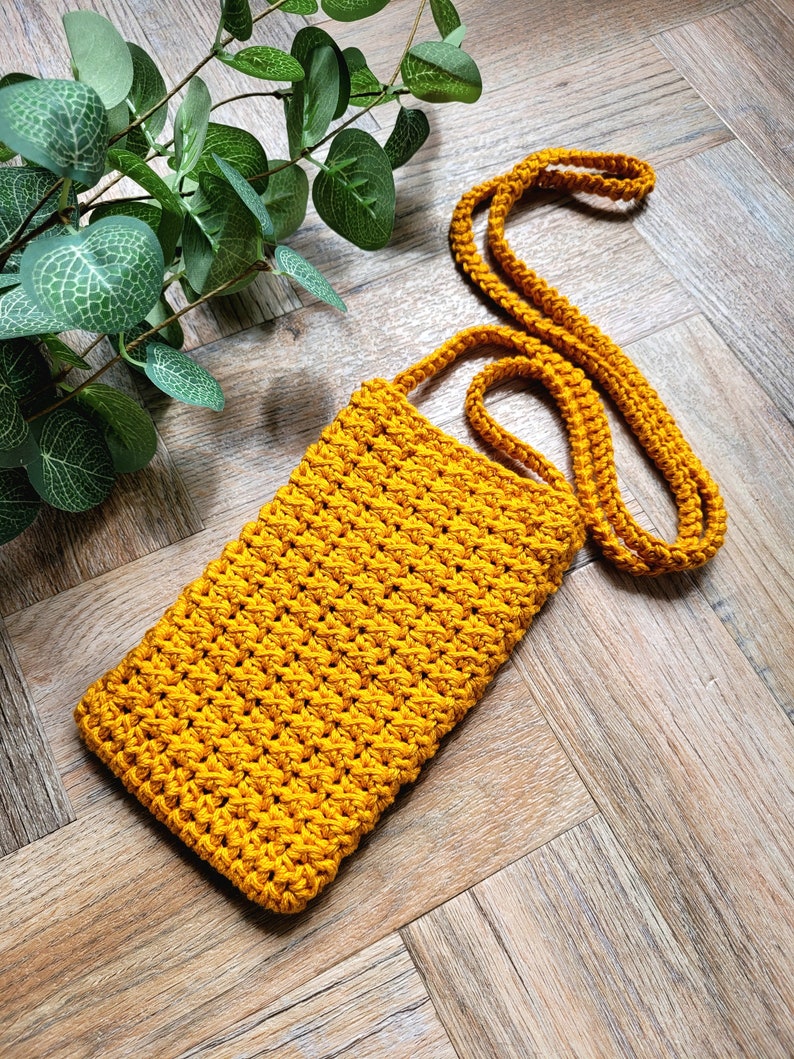 May include: A mustard yellow crocheted crossbody bag with a long strap. The bag is made of a thick yarn and has a simple, minimalist design.