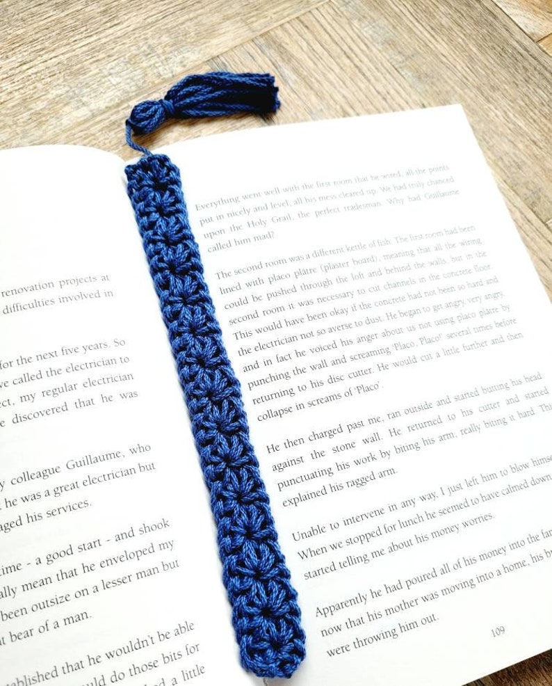 May include: A close-up of a blue crocheted bookmark with a tassel. The bookmark is lying on top of an open book with text about a renovation project.