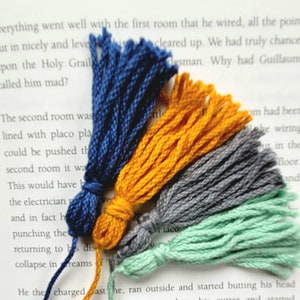 May include: Four colorful yarn tassels in blue, yellow, gray, and green. The tassels are arranged in a fan shape and are lying on a white surface.