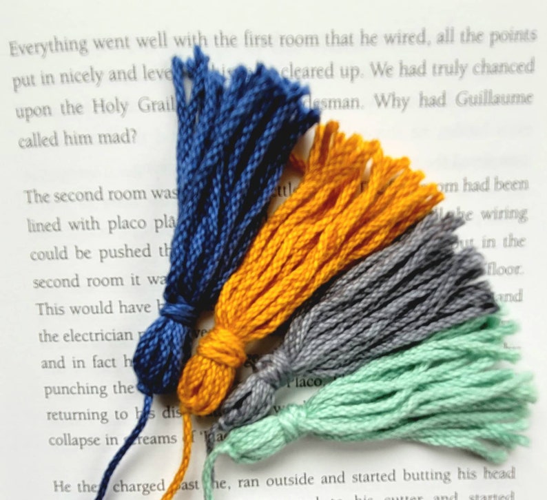 May include: Four colorful yarn tassels in blue, yellow, gray, and green. The tassels are arranged in a fan shape and are lying on a white surface.