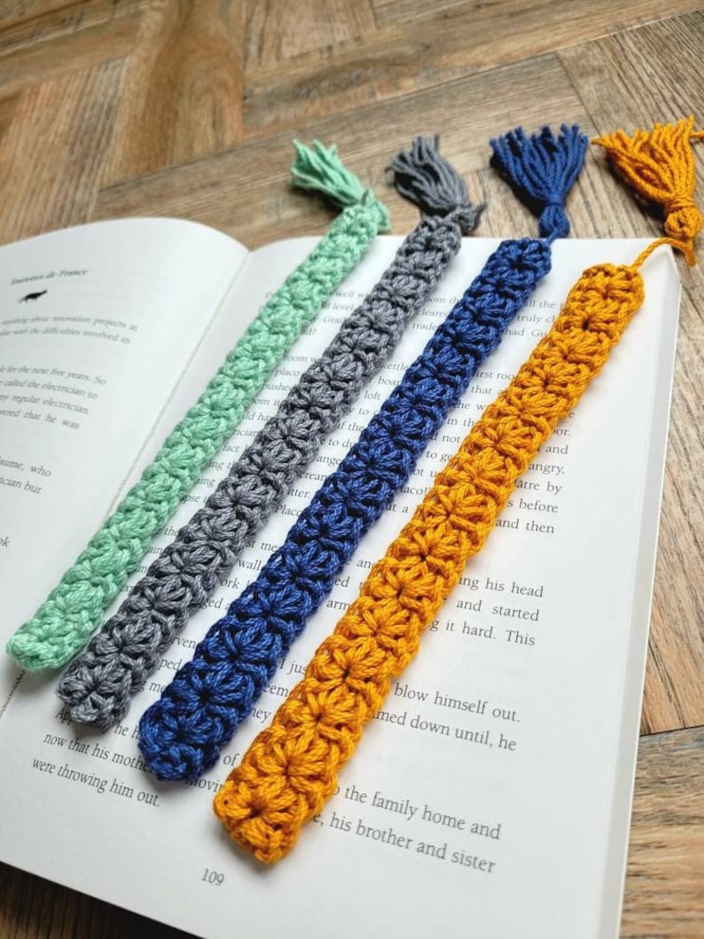 May include: Five crocheted bookmarks with tassels in different colors: mint green, gray, navy blue, mustard yellow, and a light brown. The bookmarks are lying on a book with text.