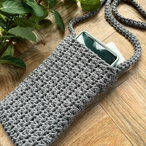 May include: A gray crocheted phone pouch with a long gray crocheted strap. The pouch is lying on a wooden surface with a smartphone inside.
