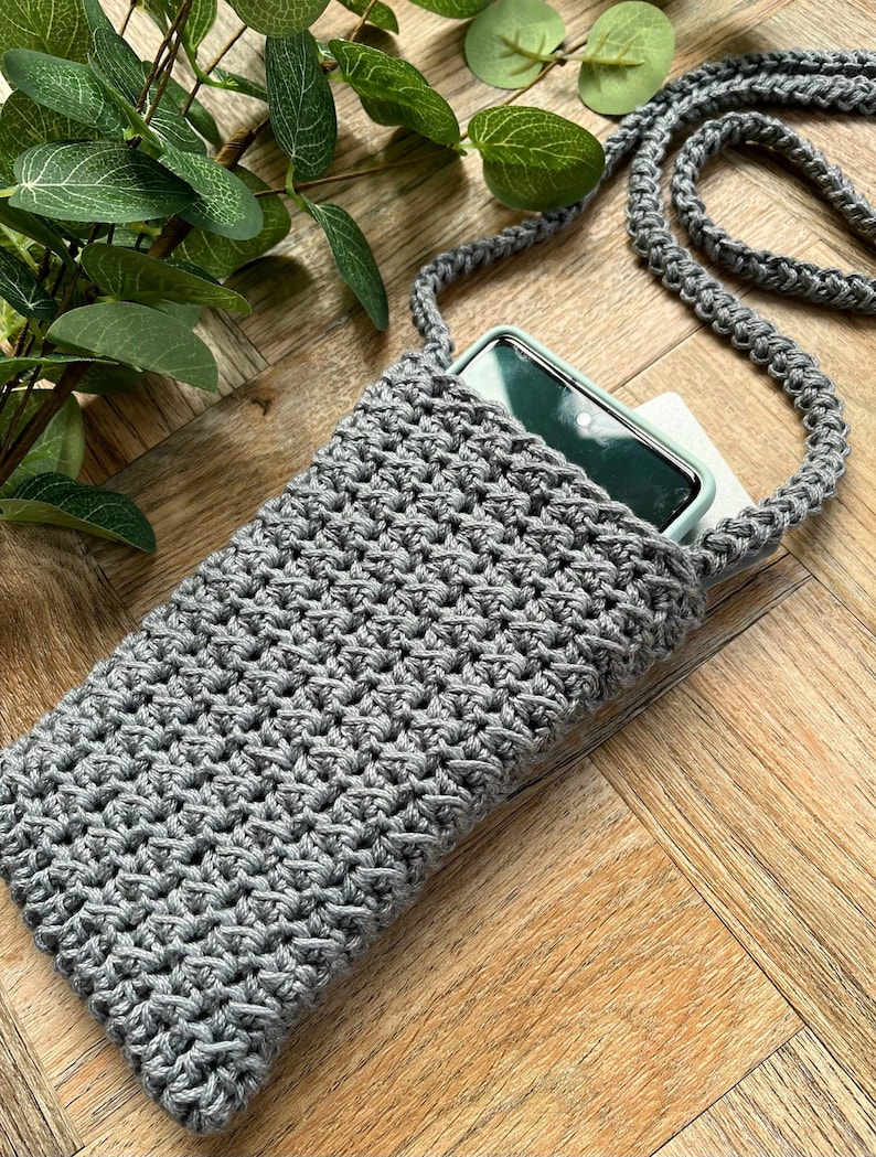 May include: A gray crocheted phone pouch with a long gray crocheted strap. The pouch is lying on a wooden surface with a smartphone inside.