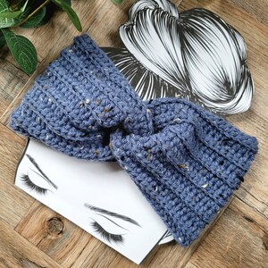 May include: A blue crocheted headband with a speckled texture. The headband is tied in a knot and is shown on a white background with a drawing of a woman's face.