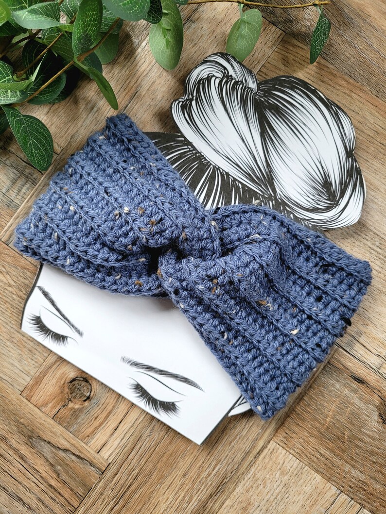 May include: A blue crocheted headband with a speckled texture. The headband is tied in a knot and is shown on a white background with a drawing of a woman's face.